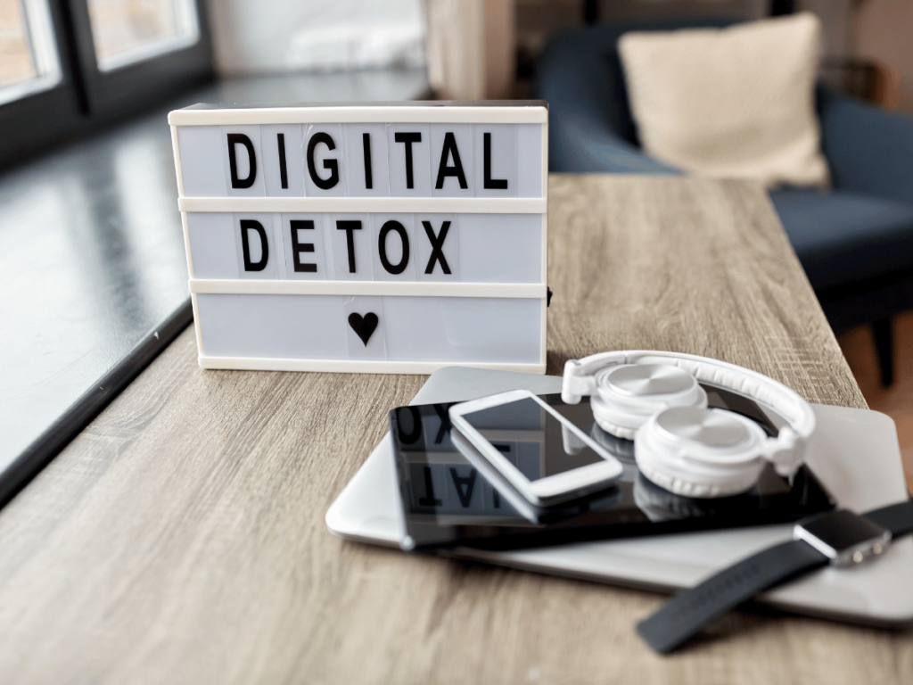 Image of a sign that says digital detox on a table with a laptop, headphones smart watch, cell phone, and tablet.
