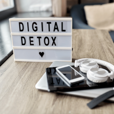 Image of a sign that says digital detox on a table with a laptop, headphones smart watch, cell phone, and tablet.