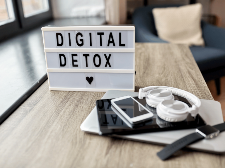 Image of a sign that says digital detox on a table with a laptop, headphones smart watch, cell phone, and tablet.