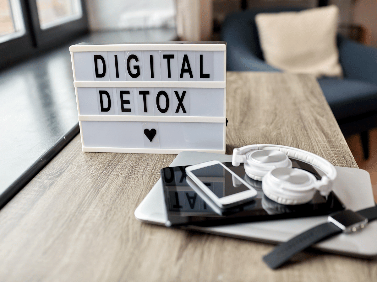 Image of a sign that says digital detox on a table with a laptop, headphones smart watch, cell phone, and tablet.