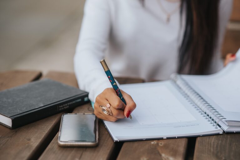 Image of a woman writing in her planner with a cell phone and book sitting next to her