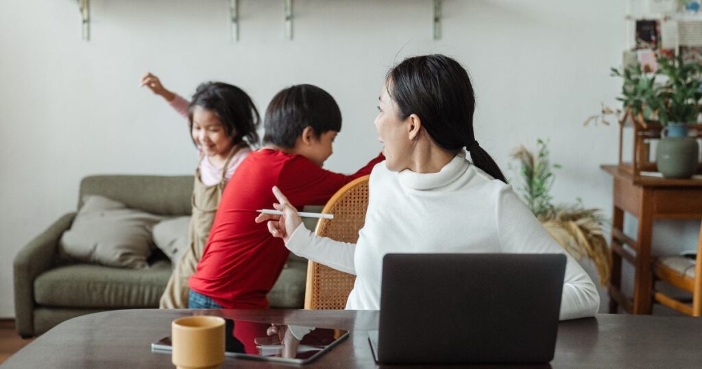 Image of a mom working at home with her kids in the background. She appears to be getting after them because she is pointing a finger at them. For a blog about work from home moms to help them balance working and caring for kids at home. 
