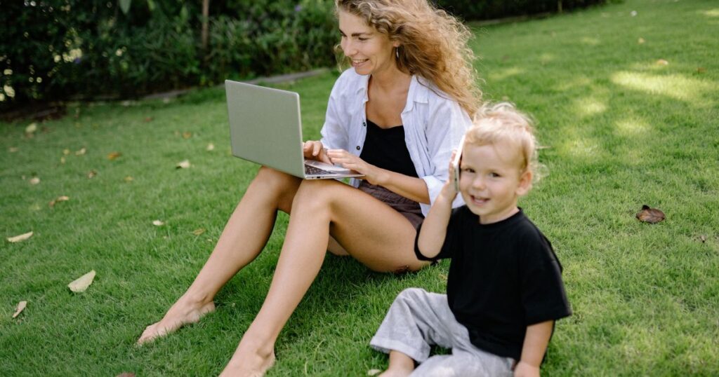 work from home mom outside on her laptop and her toddler son is next to her on a cell phone