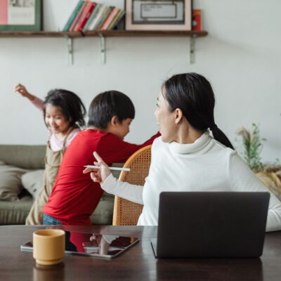 square image of a work from home mom sitting at her laptop and kids in the background playing around. SHe is point her finger at them to calm them down.